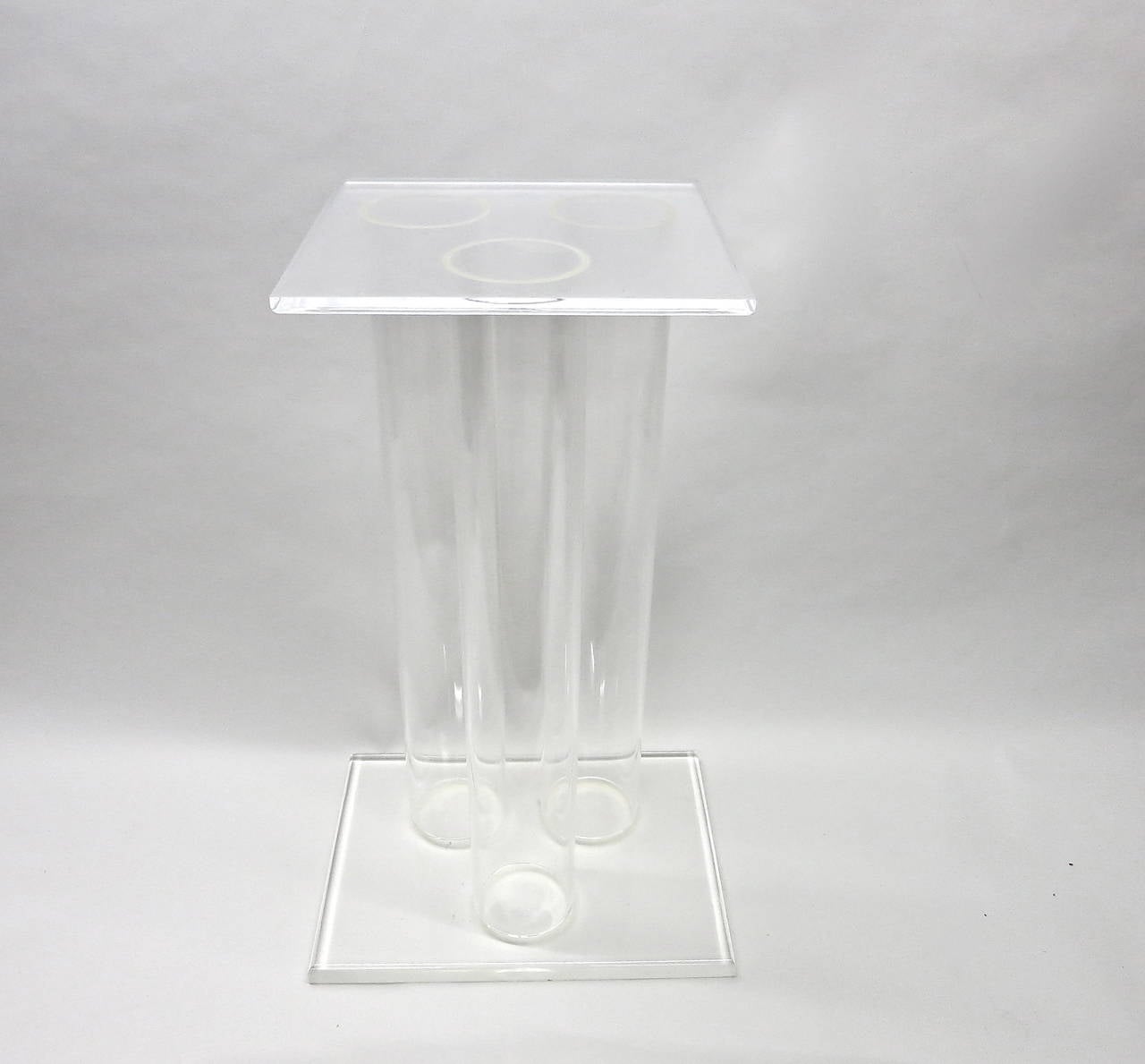 Pedestal in clear Lucite with three 5 inch diameter cylinders attached to a rectangular base positioned in a triangle and Stand vertically to support a square top.