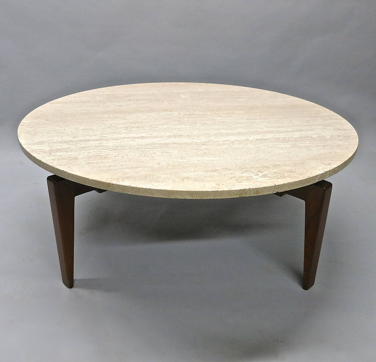 Coffee table with four walnut legs that taper from inside and all meet in the center to support a round travertine top that can lock in a set position or freely rotate continuously. Signed with a rectangular metal label applied underneath.