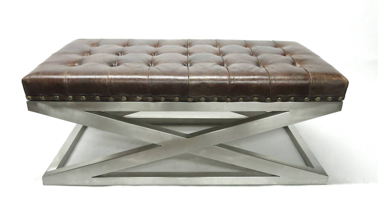 Bench has a rectangular brushed steel frame that cross in the center without any seams all supporting a brown tufted leather seat accented with brass nailhead detail.