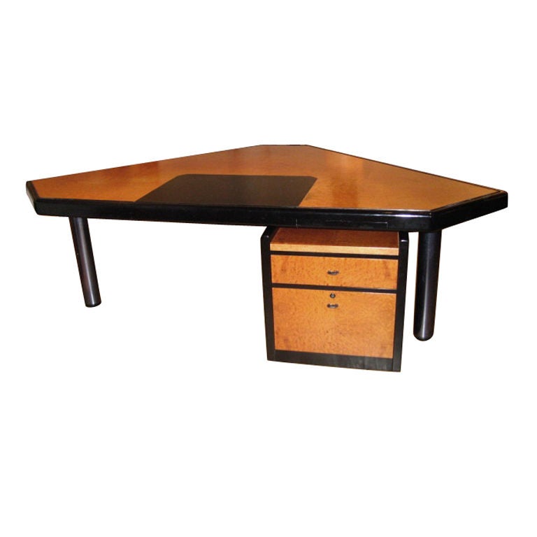 Desk is made of burled and ebonized wood supported by three enameled steel legs. The large triangular top has cropped corners and a black leather writing surface with two narrow drawers on each side. The desk has additional set drawers on casters