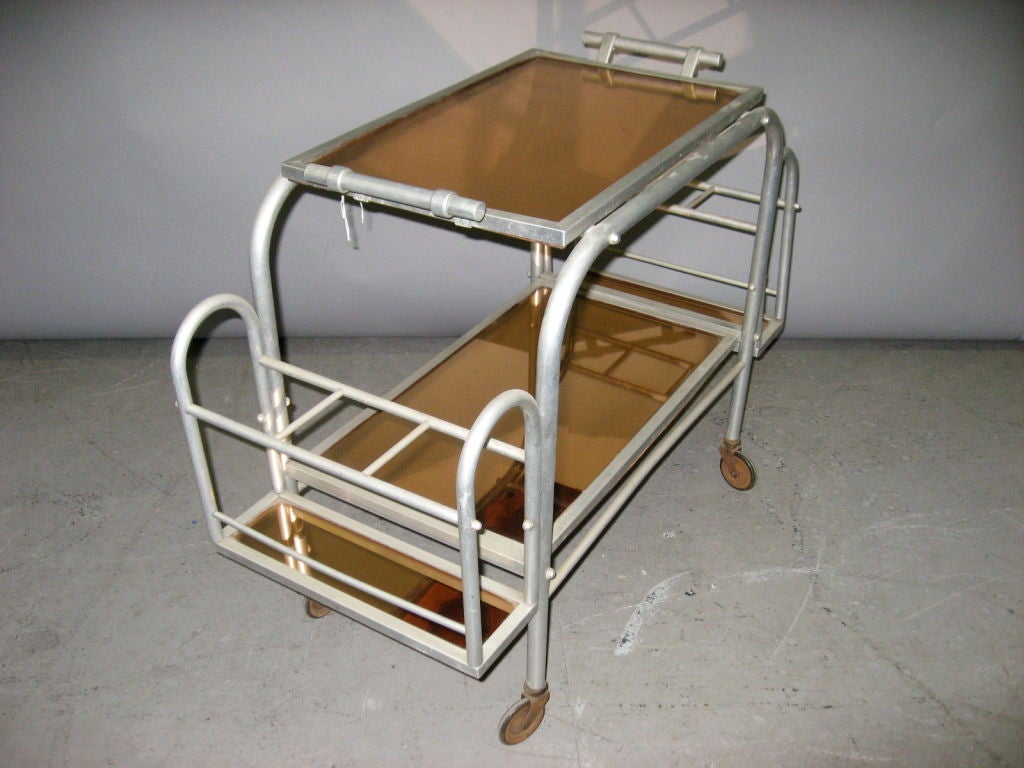 French deco bar cart made of tubular aluminum and mirrored glass. The cart has two levels with a removable serving tray on top and a fixed shelf below both in bronze-backed mirrored glass. At each end is a rack for bottle storage. Three bottles on