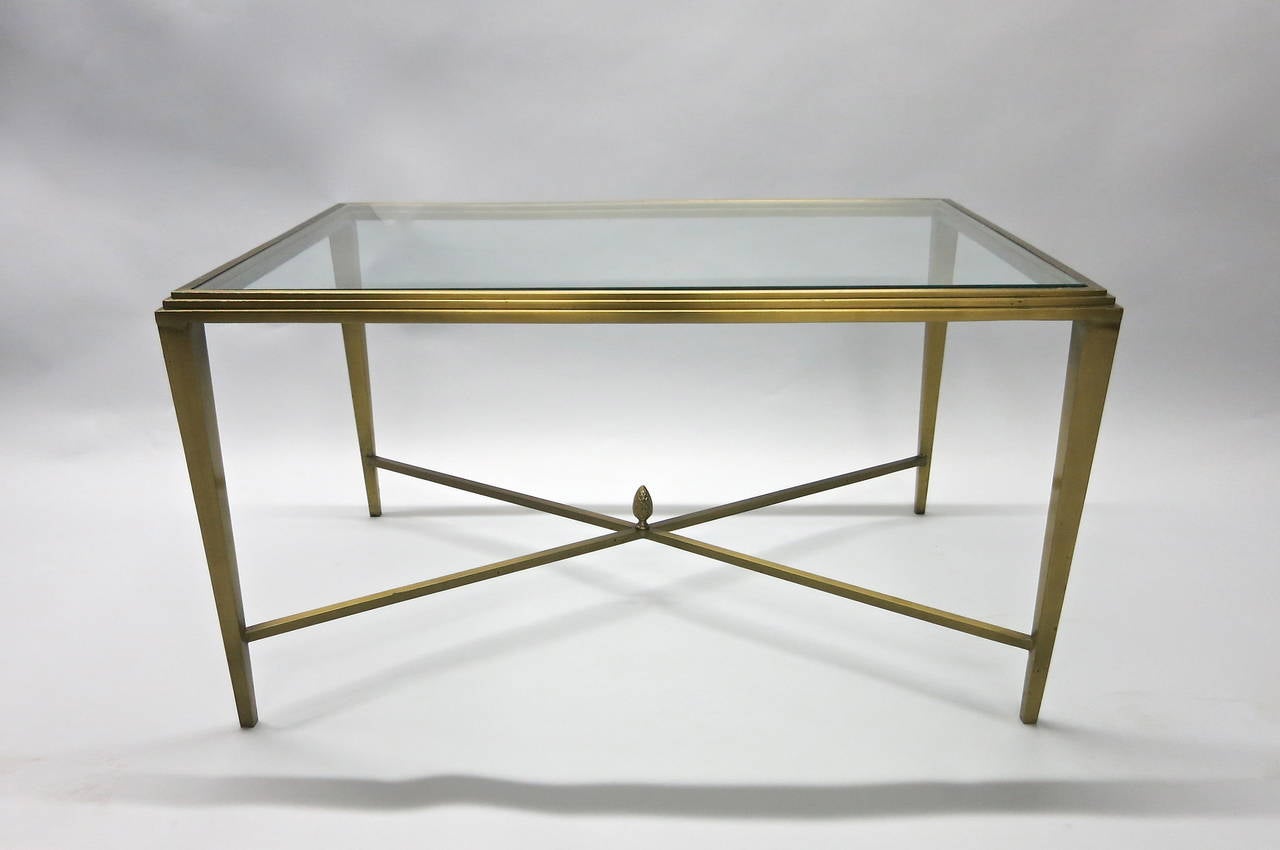 Custom commissioned coffee table in solid brass made in New York with a center inset glass top, a tri-level step detail, four tapered legs, and an x-shaped stretcher with an ovoid figure that has decorative scaling.