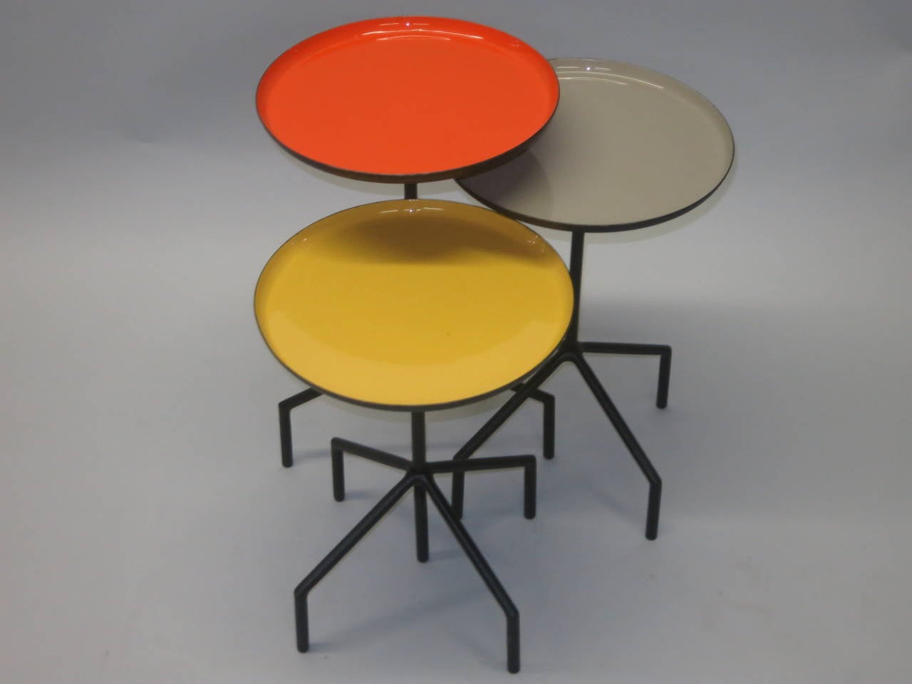 Set of three tables are composed of welded black steel rods with grey orange and yellow, round enameled, tops. The shortest is 16.5 inches high and yellow. The middle is 18.75 inches tall with a grey top. The tallest is 21inches with an orange top.