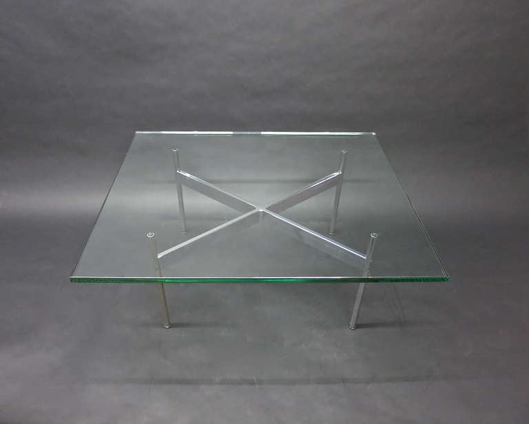 Coffee table in polished metal with a glass top that sits on a rectangular base with X-shaped stretcher and four round legs each with rubber supports at top and adjustable feet at bottom.