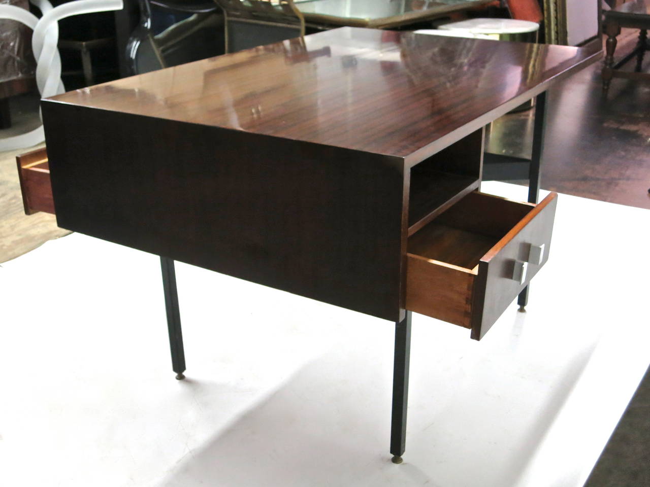 Asymmetrical partners desk in mahogany wood with one shelf and one drawer on each side, all supported on a metal base that has a stretcher with ends bent to two inward facing triangular forms, and four solid brass and adjustable feet. Each drawer