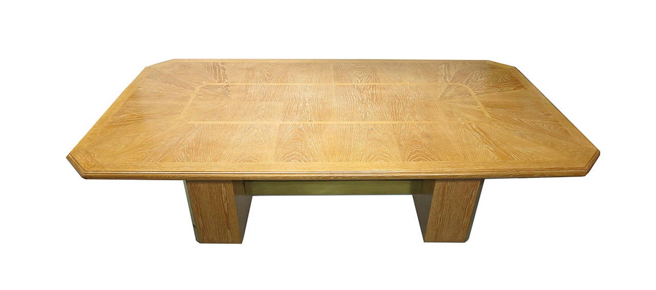 Strong dining table in limed oak with a long, six-sided top and brass detail in the base. The top has rounded and tri-level, routed edges with a subtle sunburst pattern of triangles at each end that borders the rectangularly joined wood in the