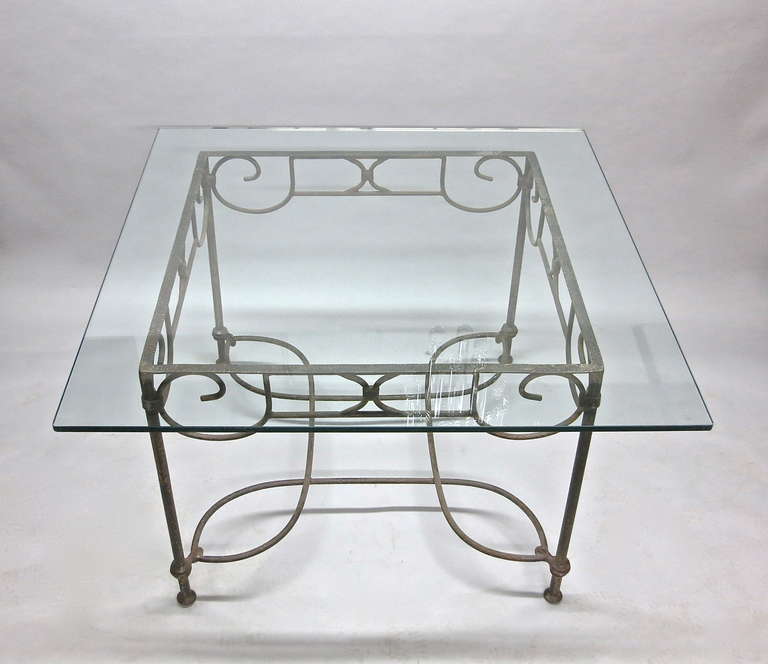 Square table, with a clear glass top, in patinated wrought iron with decorative detail on each side and an arched stretcher at the bottom that connects the legs.
