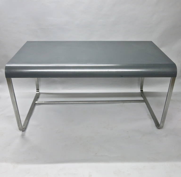 Desk in metal with brushed steel legs on each side connected by a off centered stretcher at the floor. The top is enameled grey and arched front and back.