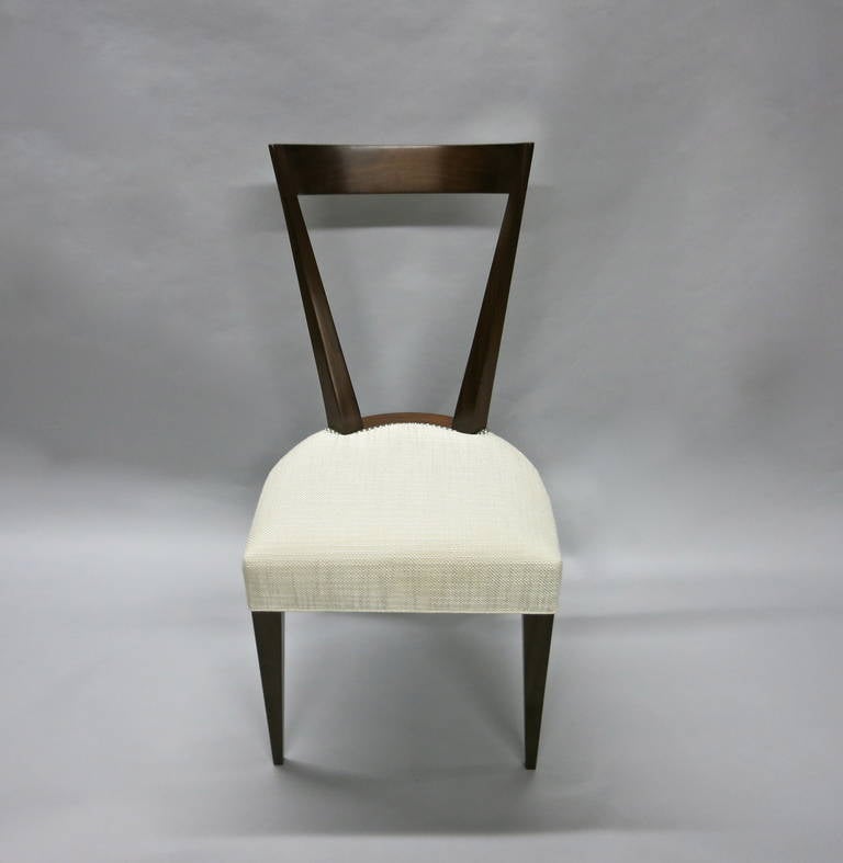 Dining chairs in dark stained wood with an angular, open and arched back, an upholstered seat trimmed with nail heads resting on four tapered legs.
