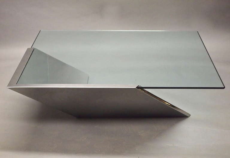 'SMT Table' by Brueton has a 1/2 inch reverse bevel, tempered glass top that is 48 inches square. The base is 1” thick stainless steel panels truncated inward at a 45° angle to form a L-shaped base; welded, ground and polished into a seamless unit.