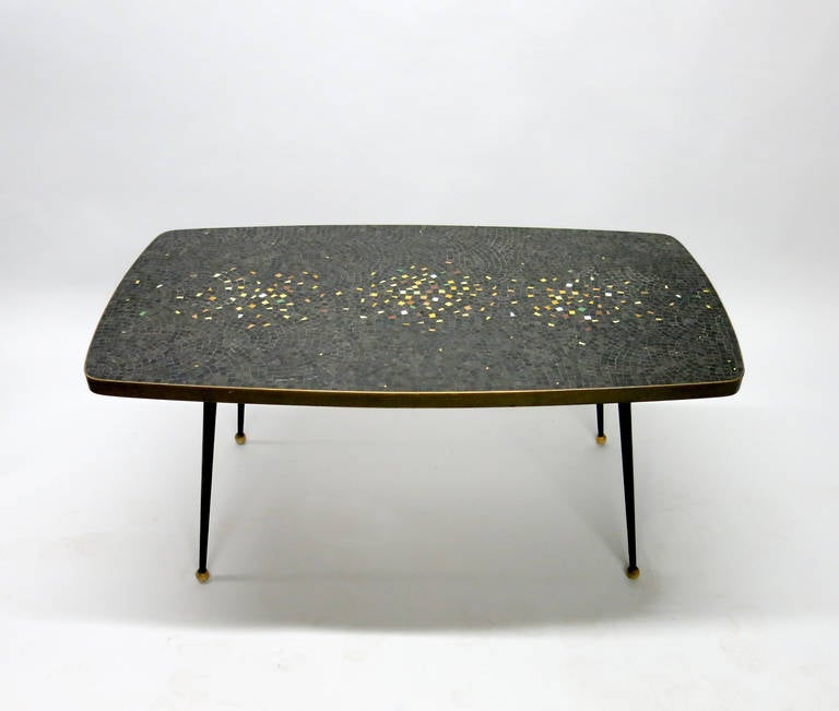 Coffee table with a top made of inset mosaic tile that is predominantly black accented with a great multi-color tile scatter detail framed with a solid, patinated brass border. The table's four legs are tapered, black enameled metal and have round