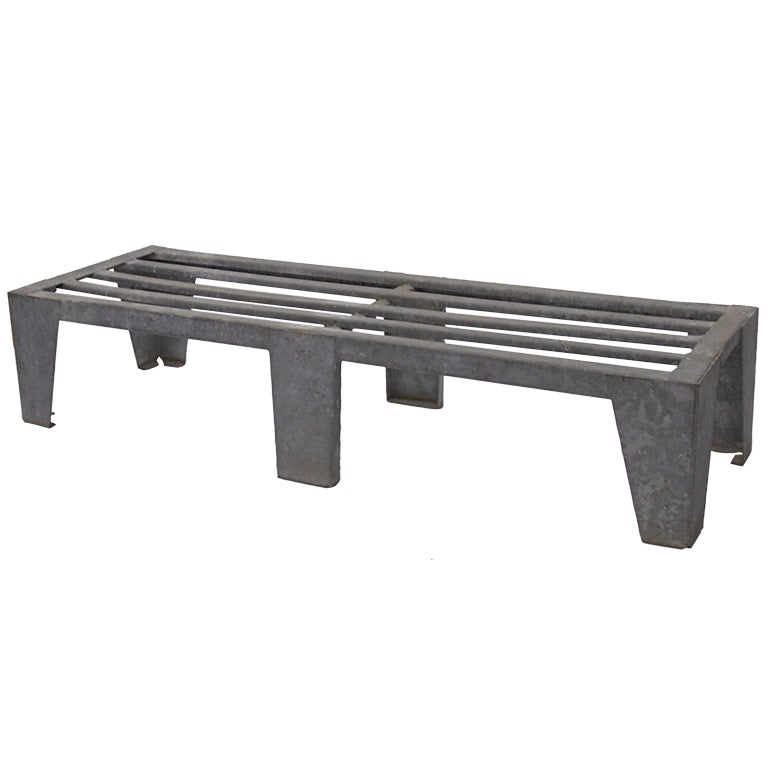 Industrial bench in galvanized steel with four corner legs that taper, two center legs that are rectangular and support a cross stretcher. The top consists of galvanized steel bands that run the length of the bench and are spaced to endure the