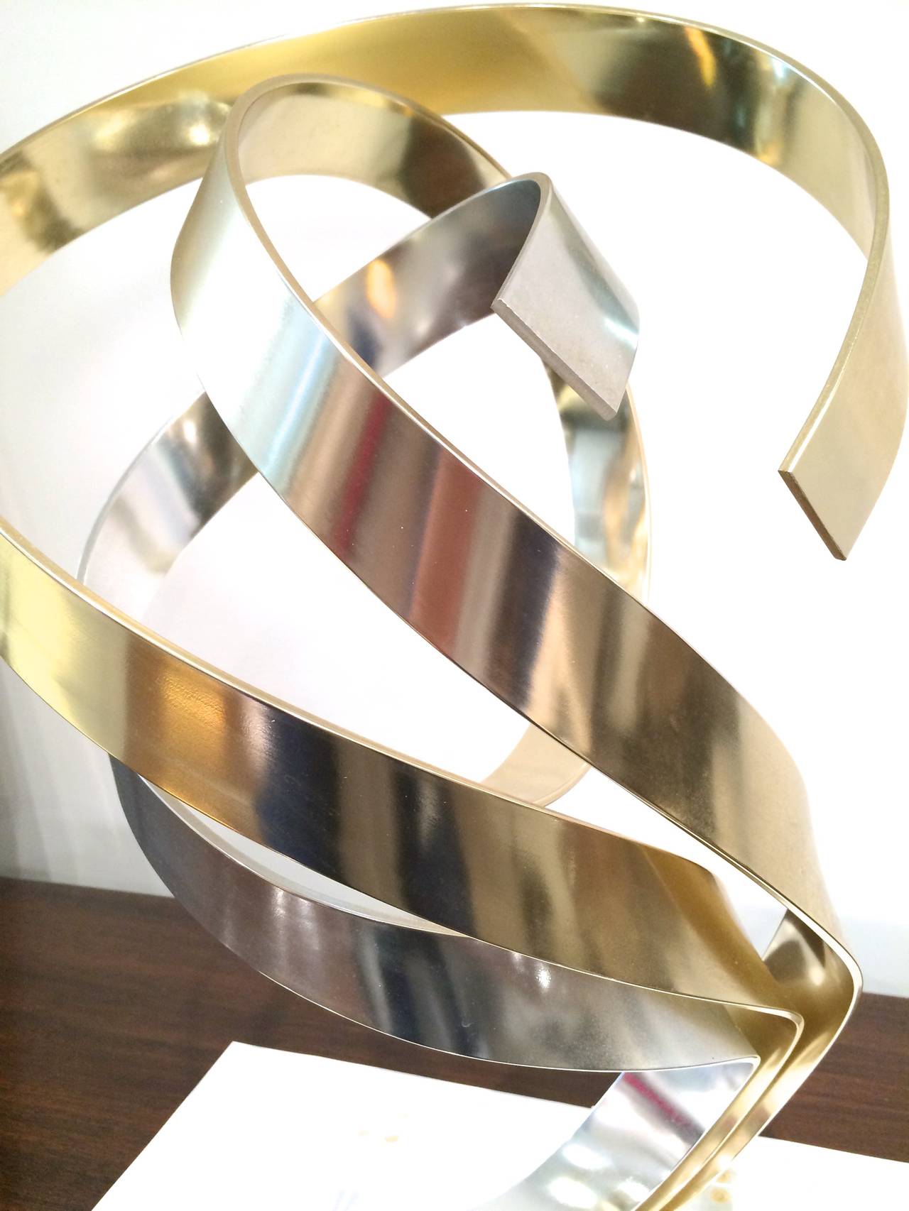 Signed triple ribbon sculpture by Dan Murphy, circa 1987. Gleaming interlocking swirls of anodized silver and gold tone aluminum are woven together atop a solid Lucite base.