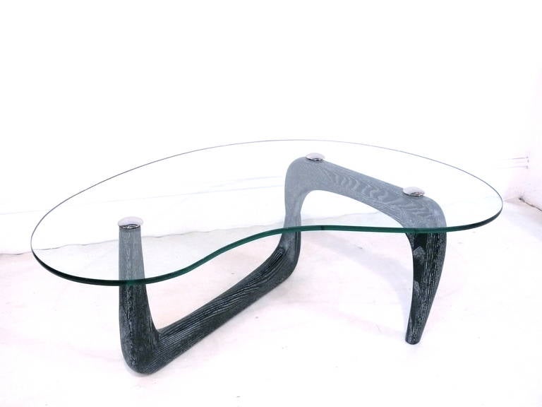 Gorgeous biomorphic glass top coffee table in the manner of Gilbert Rohde. The free-form glass is fastened with three nickel-plated caps to its beautiful oak base refinished in black ceruse.