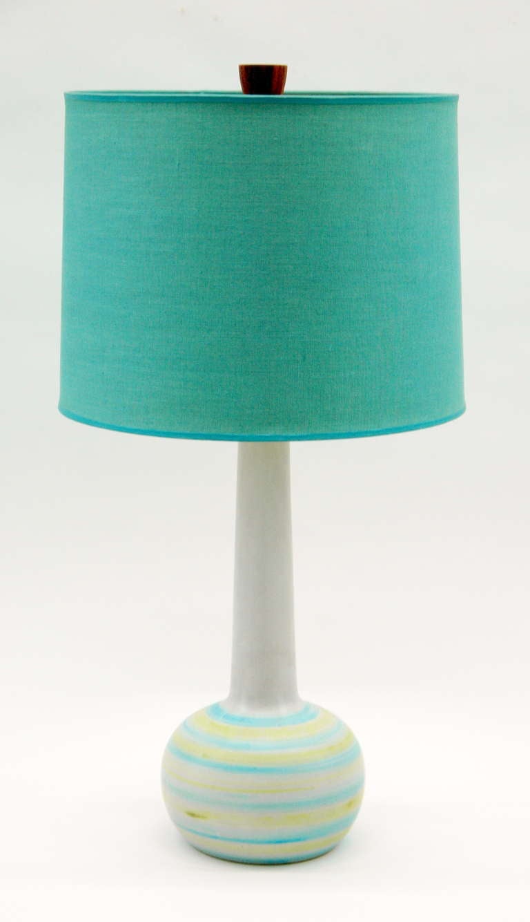 A ceramic table lamp by Gordon and and Jane Martz with a spherical base and elongated neck. The lamp has a white finish with green and blue stripes irregularly painted on the lower part. The blue linen lamp shade and oak finial are original  to the