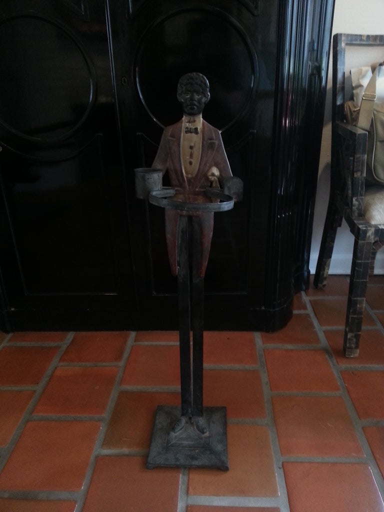 A figural umbrella stand or cane holder in cast, painted metal. The black valet dressed in bowtie and burgundy tails, his lower body acting as pedestal atop a square platform. 

Early 20th century, United States. Model number 136 impressed on