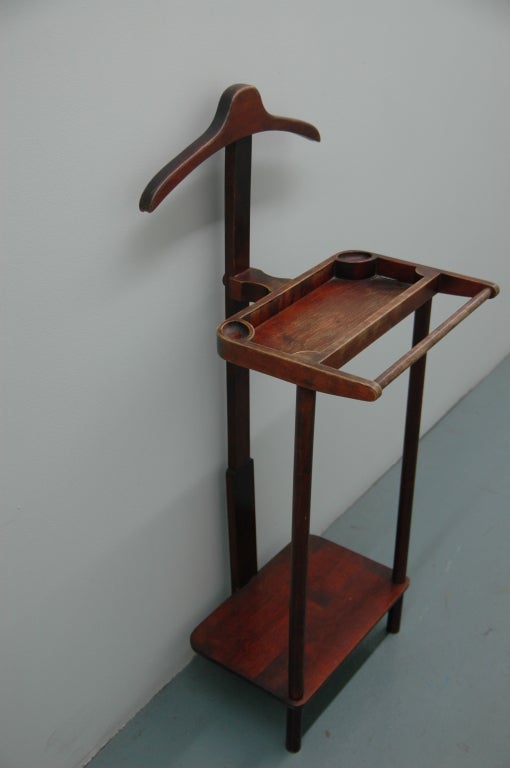 A vintage wooden valet stand stained in mahogany with a segmented tray with a pants bar at front and a shoe shelf below surmounted by a hanger hook.