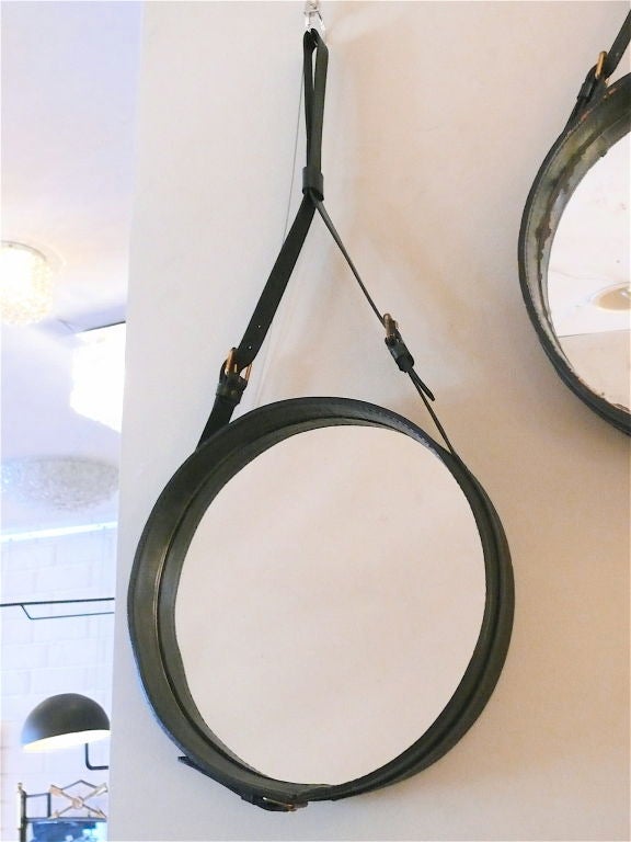 Original black leather mirror by Jacques Adnet, the round inset mirror hanging by a leather strap with brass buckle detailing. Two available.
