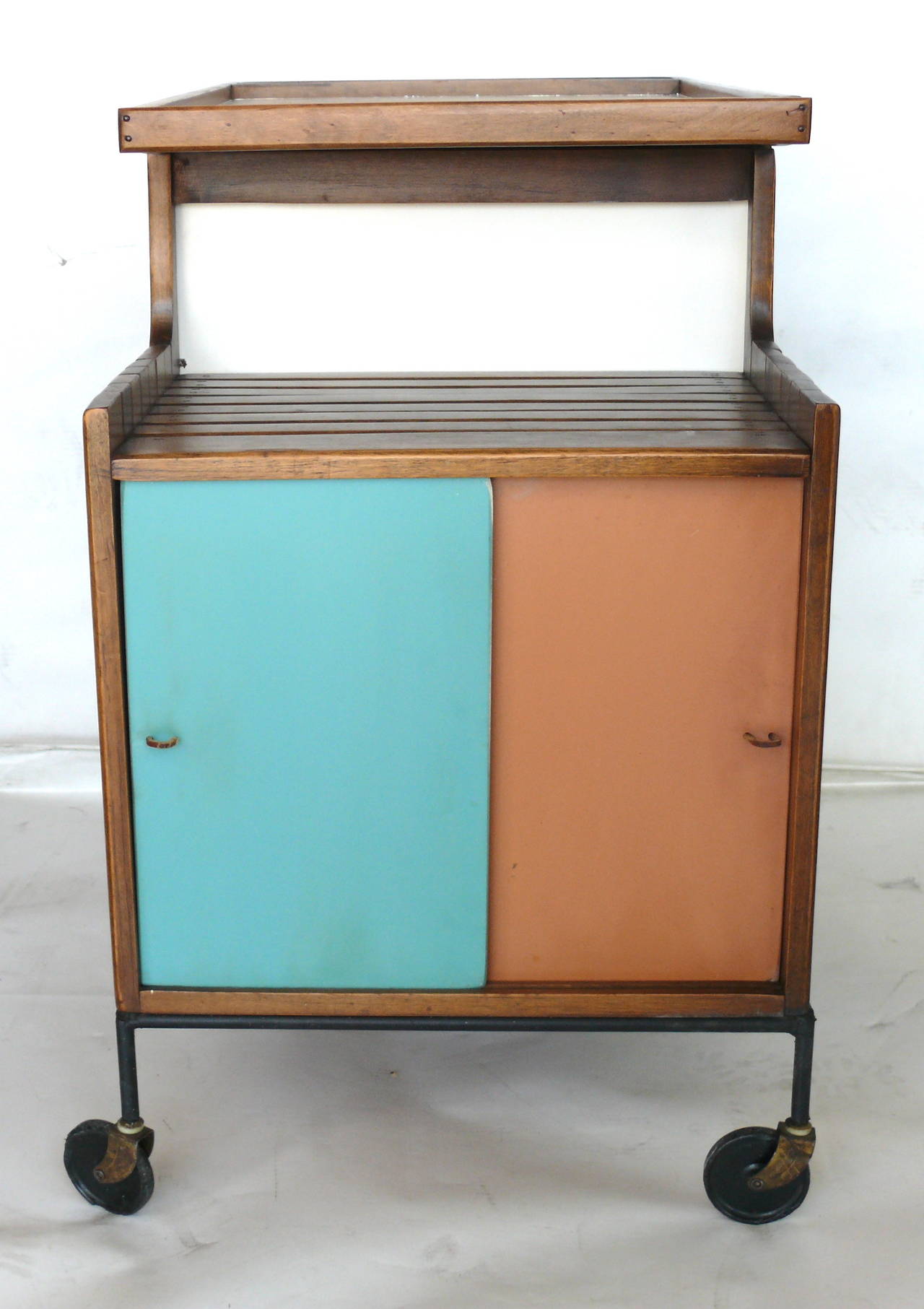 Whimsical bar cart by Arthur Umanoff on brass casters. Iron framed cart features slatted wood, a tray top, multiple shelves and an enclosed cabinet. Sliding doors have two different colors that you can reverse and interchange. Great colors and in