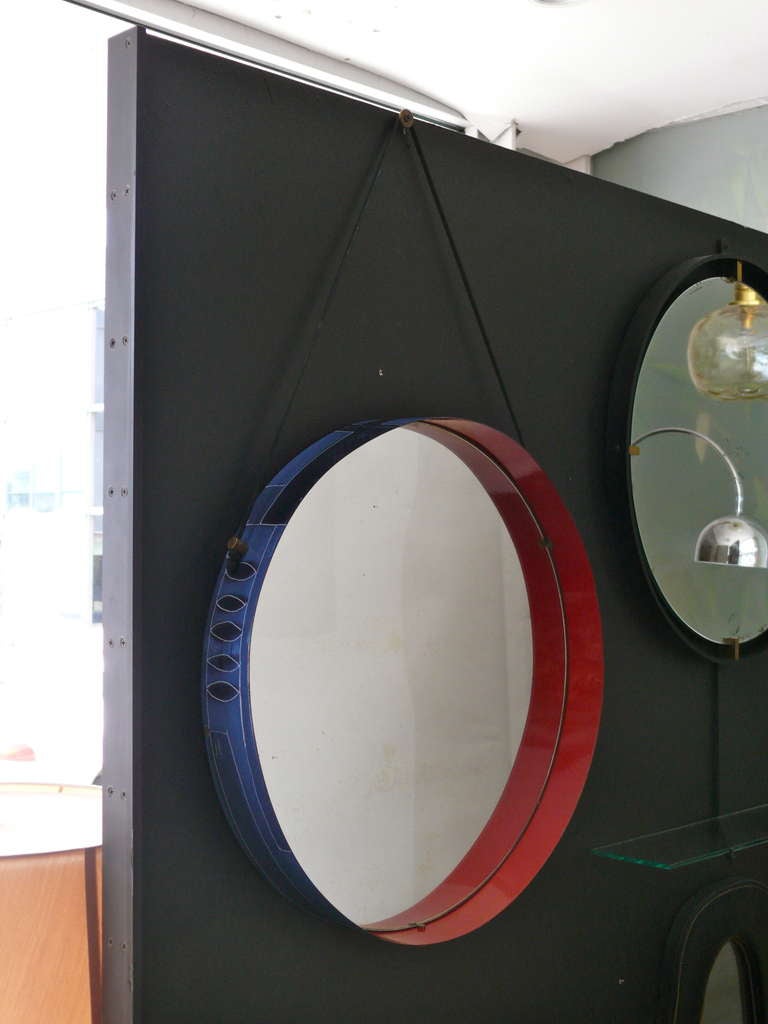 Stunning Italian circular wall mirror in blue and red ceramic on metal frame. Exterior of frame has painted shades of blue with leaf and geometric design. Interior is painted in a deep red lacquer. New black leather strap connects at two original