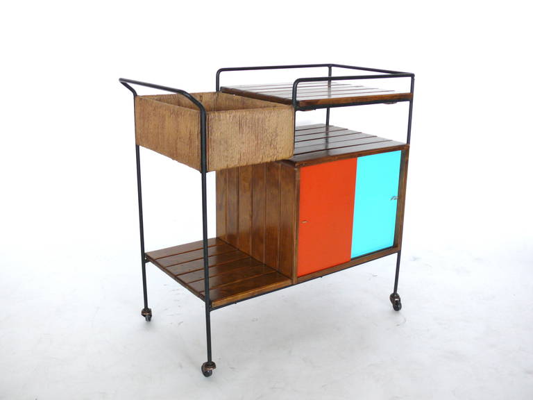 Whimsical bar cart by Arthur Umanoff on wheels. Iron framed cart features slatted wood, a tray top, a rattan inset tray, and an enclosed double sided cabinet with red and blue cabinet doors. Great colors and in excellent vintage condition.