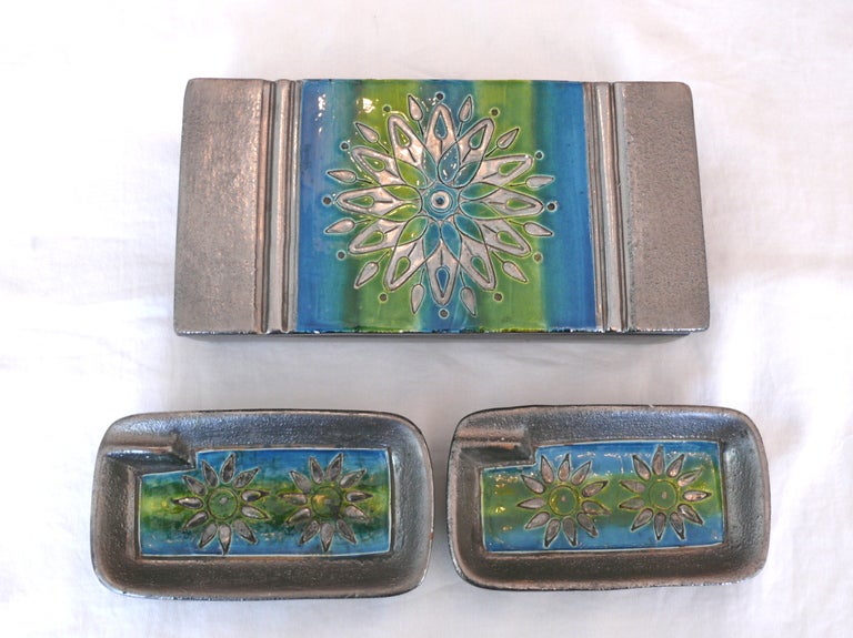 Striking set of Italian ceramic cigarette box and two small ashtrays. Painted in silver, green and blue with etched flowers. Beautiful set of objects to display or perfect as a gift. Signed on the bottom of the box. Sold as a set. 
Box Dimensions: