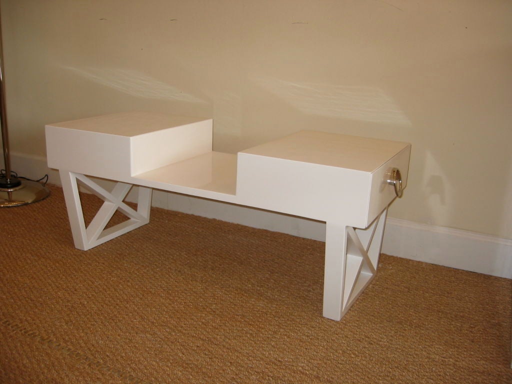 This Mid-Century Modern cocktail table is fashioned in wood and lacquered in white high gloss. The legs have inverted frames with ‘X’ centers and the table has two identical sides each with drawers with original nickel silvered hoop pulls. This is