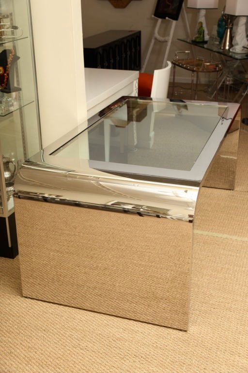 This fabulous oversized desk features two continuos waterfall sides in highly polished steel.  A thick slab of clear glass is inset in the top creating a light and airy feel. It could easily float in a room or be anchored by double sided seating. 