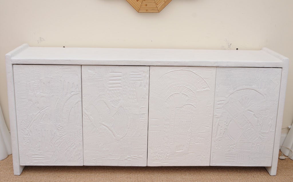 The four doors of this interesting textural and dimensional cabinet/console/buffet have designs of embossed and incised plaster of paris abstract forms; this has all been redone  and refreshed in white lacquer. It is signed on the lower left door;
