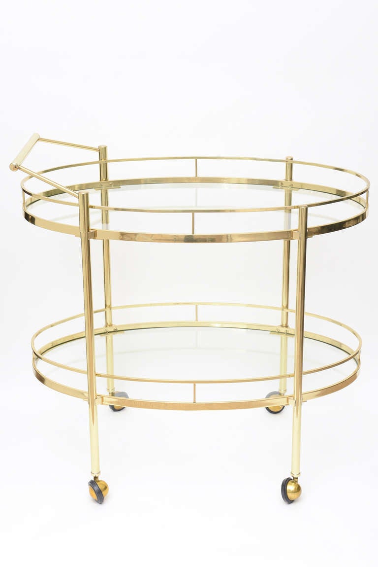 This wonderful vintage mid century modern two-tiered oval polished brass bar and serving cart /trolley is simple and elegant.
It has two glass shelves and brass handle to push. The split wheels are brass and rubber so they are perfect for marble,