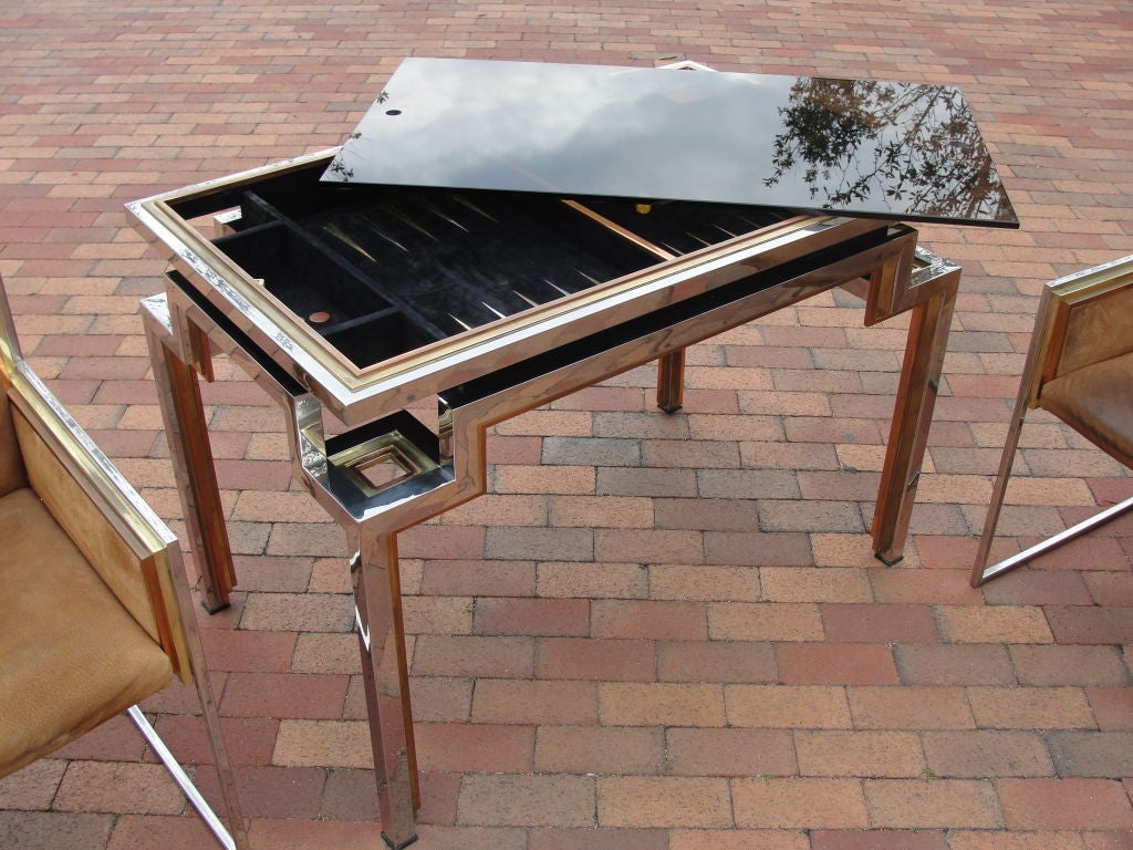 Alain Delon pour la collection Jansen - Fabriqué en 1972.  Table de backgammon en laiton, chrome et acier et paire de fauteuils assortis avec tissu original en cuir/suède.  Le plateau en verre fumé est doté de deux trous pour les doigts permettant