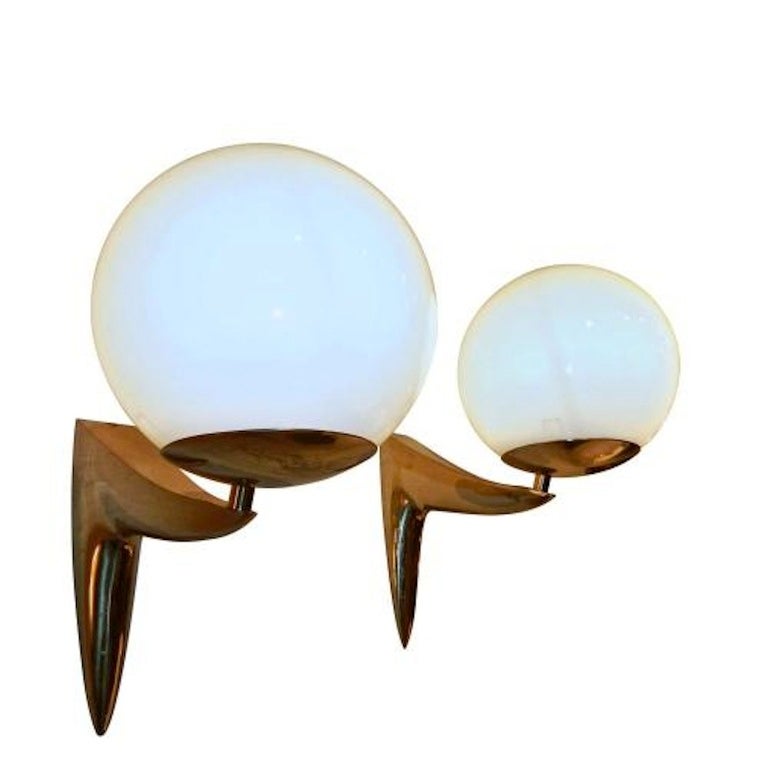 A pair of mid century wall sconces each featuring simple curving inverted L shaped bodies with pointed detailing and a shallow bowl shaped shade support which holds a white opaque glass light diffusing ball shaped shade with a top center opening.