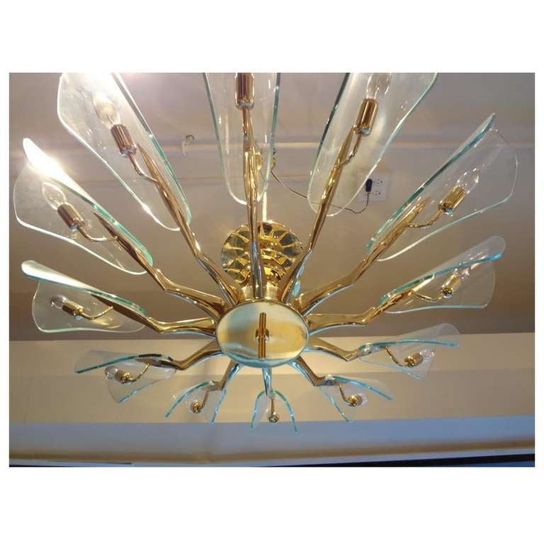 Pair of Mid-Century chandeliers each featuring a body in gilt brass with twelve shaped arms with four central disks decorating the base of the hanging rod. Each of the arms terminates in a single light source with a hood shaped shade in clear hand
