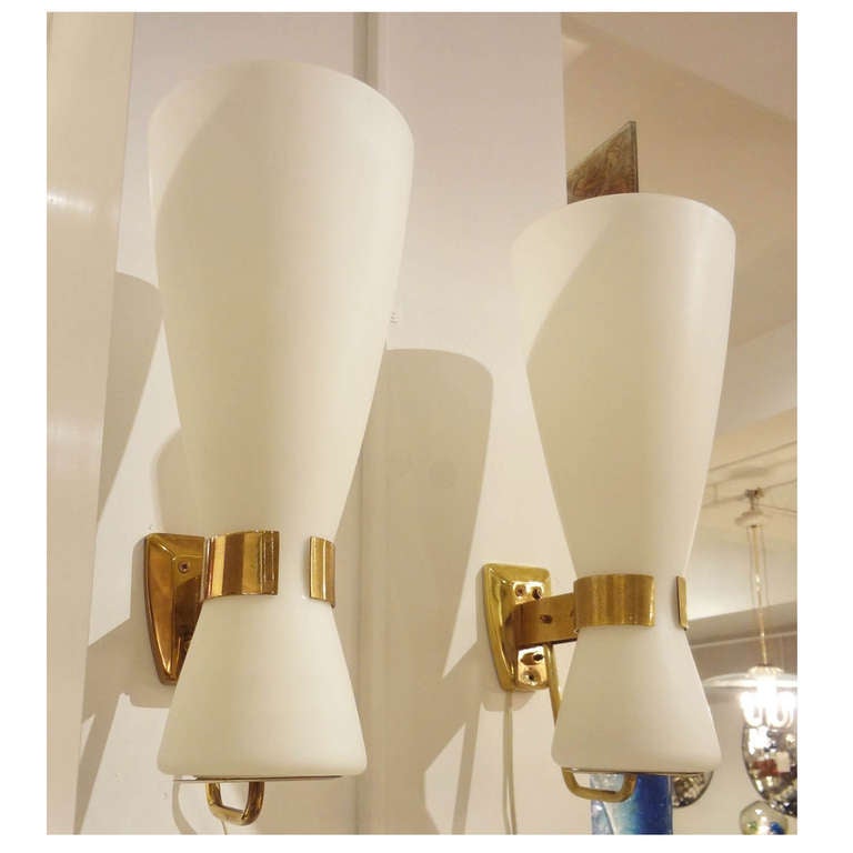 A pair of grand scaled wall sconces each featuring frames and wall brackets in gilt brass which hold large one piece white opaque light diffusing glass shades. The sconces are signed on the tops of their wall brackets, Stilnovo, Italy, circa 1959.