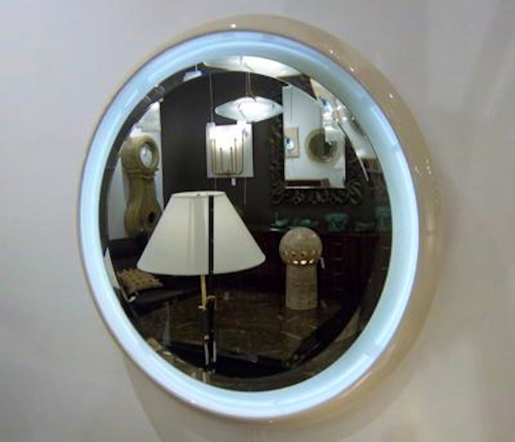 A round wall mirror featuring a inner floating clear mirror with a beveled edge which is lit from behind. The mirror also features a bowl shaped ceramic frame which has been glazed in a neutral cream color, France, circa 1965.