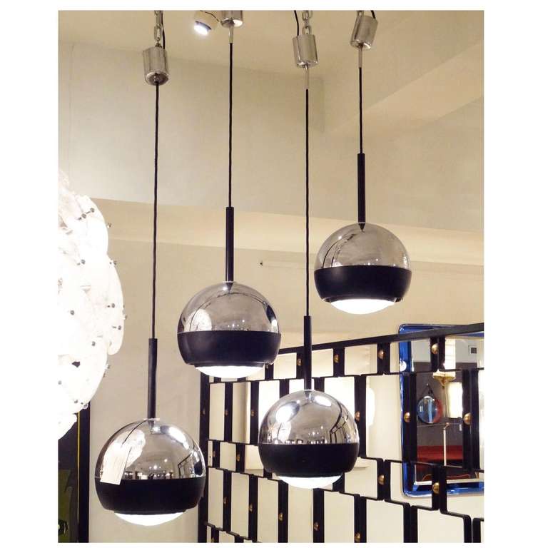 A set of four modernist hanging lamps each featuring a black cord hanging system with a ball shaped body with a chrome top half, a black lacquered steel section below the chrome and a faceted jewel-like lens which directs the light down and out,