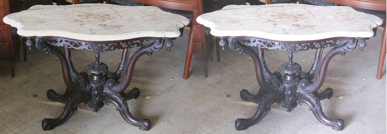 Pair of Anglo-Indian 19th Century Carved Mahogany Center Tables with Carrara Marble Tops. The shaped molded marble tops over a pierced and carved apron. The tops supported by upright and down swept supports carved with scrolls, foliage and