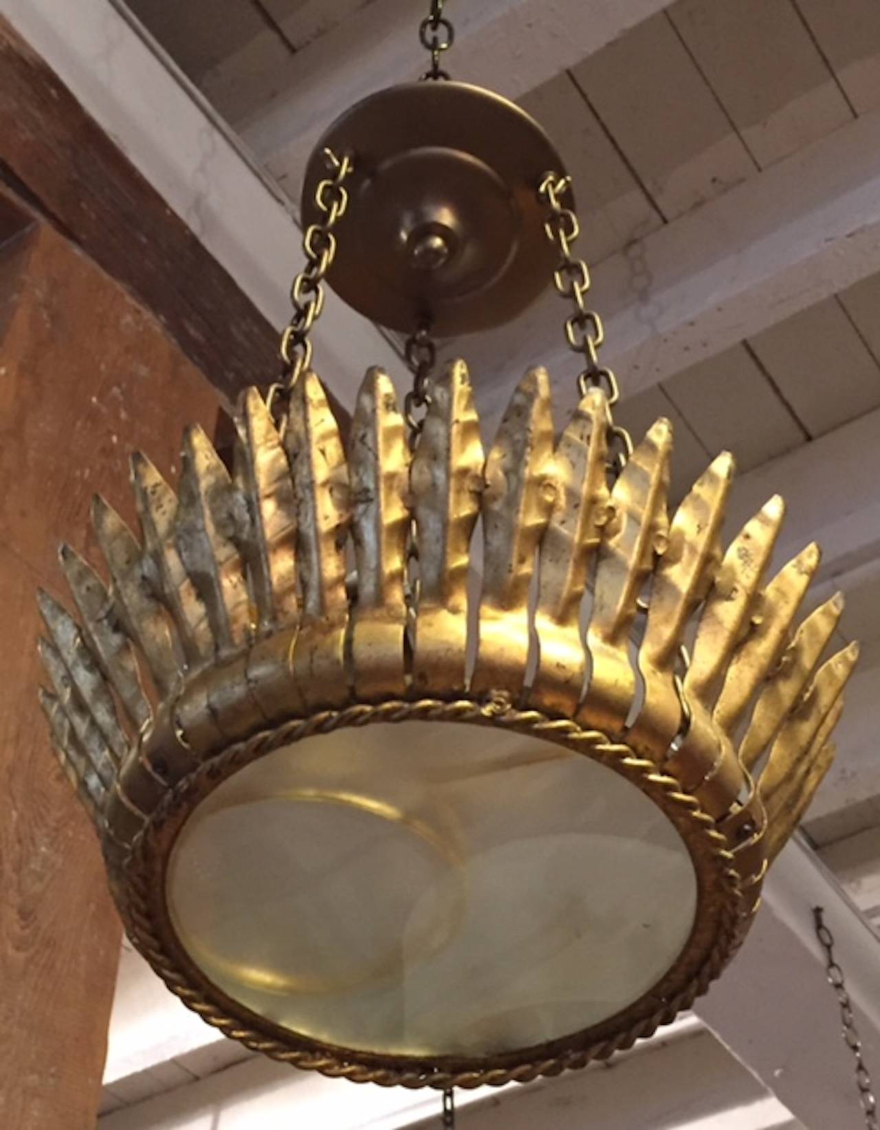 1940s Spanish gold gild metal chandelier that resembles a crown.
Gold gilt metal feathers create the crown with a gold gilt twist surrounding the rim. A smoked glass disc at the bottom of the pendant light hides the bulbs inside.
Three chains are