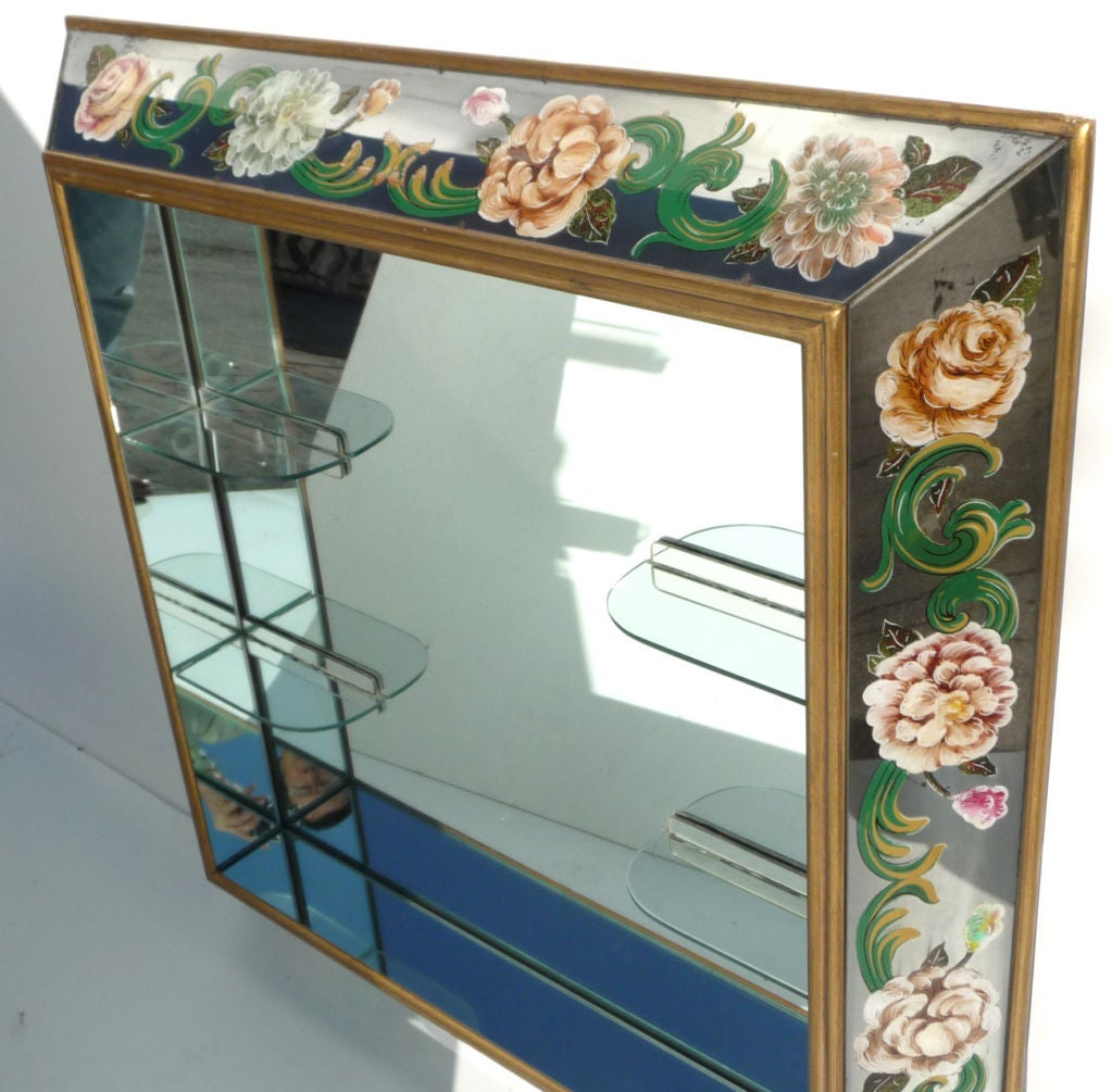 This fantastic mirror is framed in eglomise (reverse painted on glass) panels, with a repeating floral motif. The interior mirror is intersected with curved glass shelves. The entire unit is framed in a gold painted wood, with solid hanging clips on