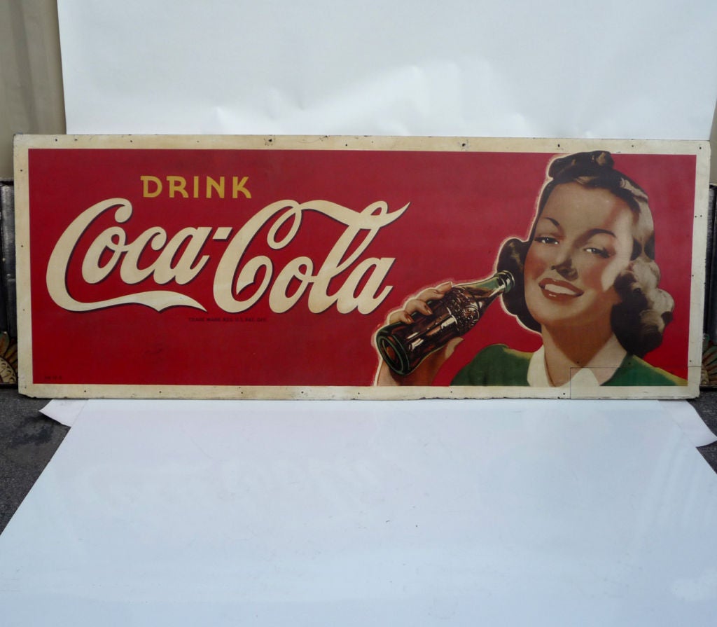 A great graphic in the classic, wholesome style favored by the worlds most popular drink. The rosy cheeked beauty beams as she prepares to enjoy her first sip of a Coke. The advertisement is screened onto a Masonite board, as opposed to the smaller