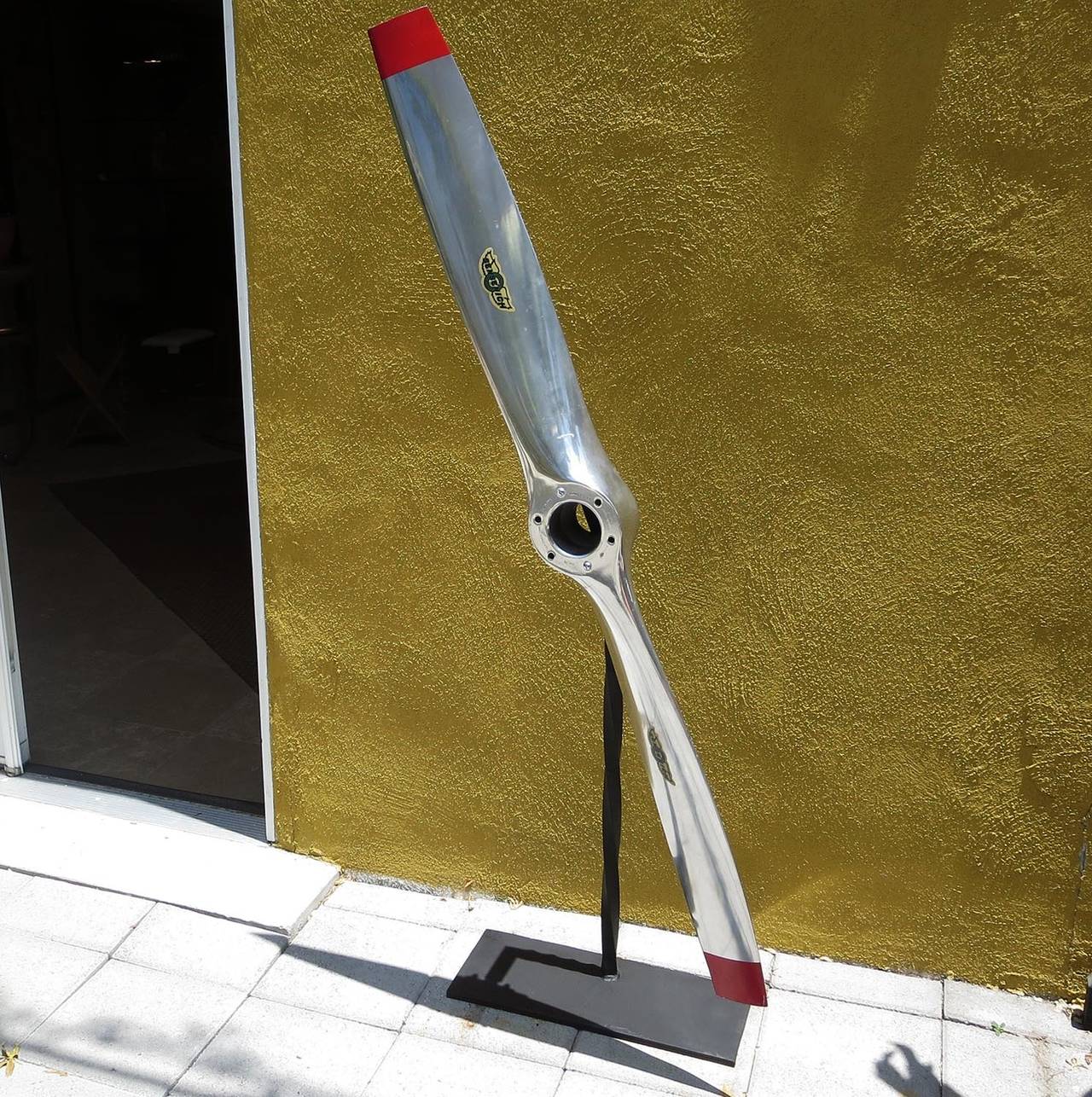 A Sensenich cast and polished aluminum propellor, complete with original decals and painted tips. The sculptural propellor free spins on a painted steel floor Stand. The length of the prop is 64 1/2 inches, although the overall height is taller,