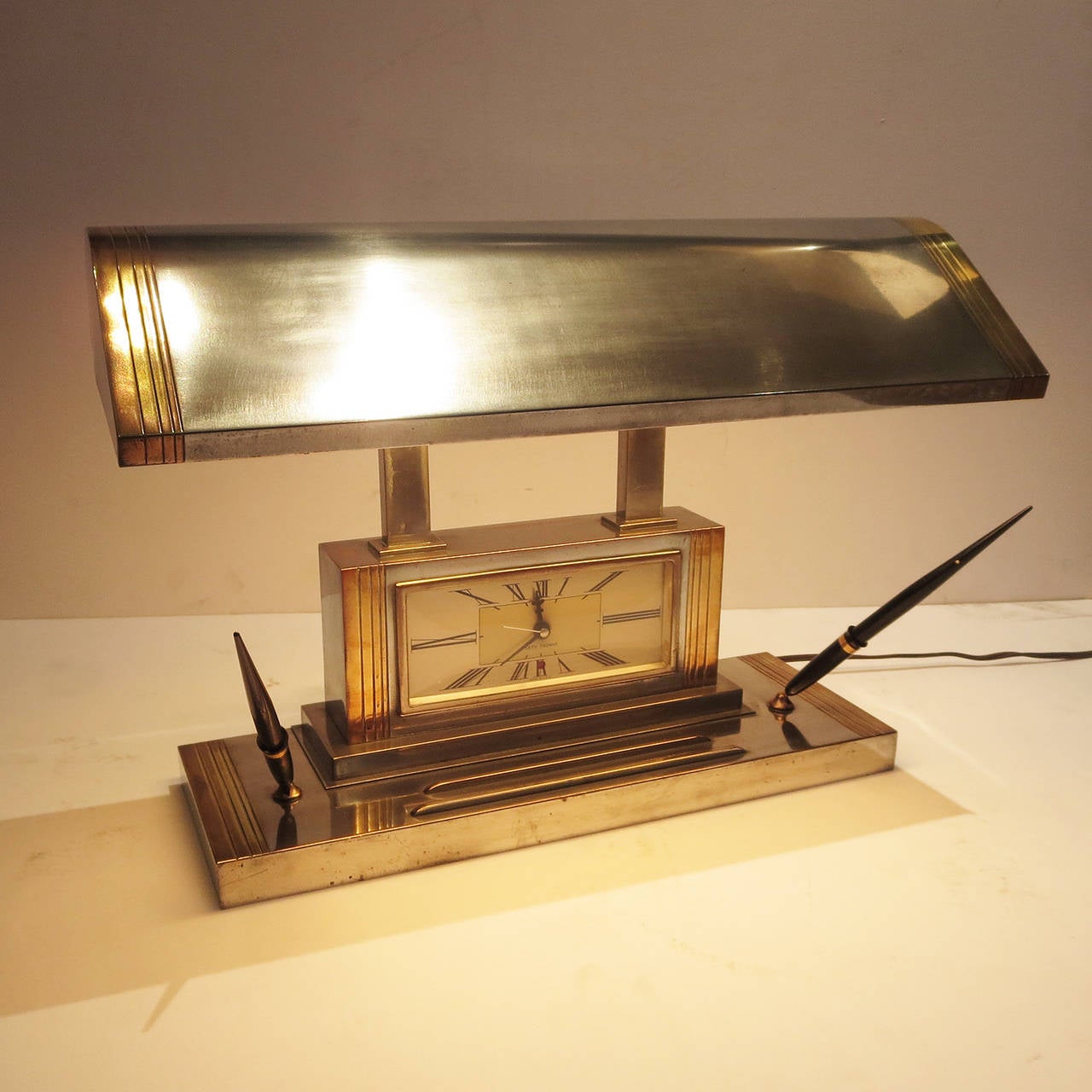This stylized desk lamp is in perfect working condition, including the clock mechanism. They are usually found without the pair of pen holders, making this version a bit more unusual. The lamp is a combination of silvered and bronzed metals, and