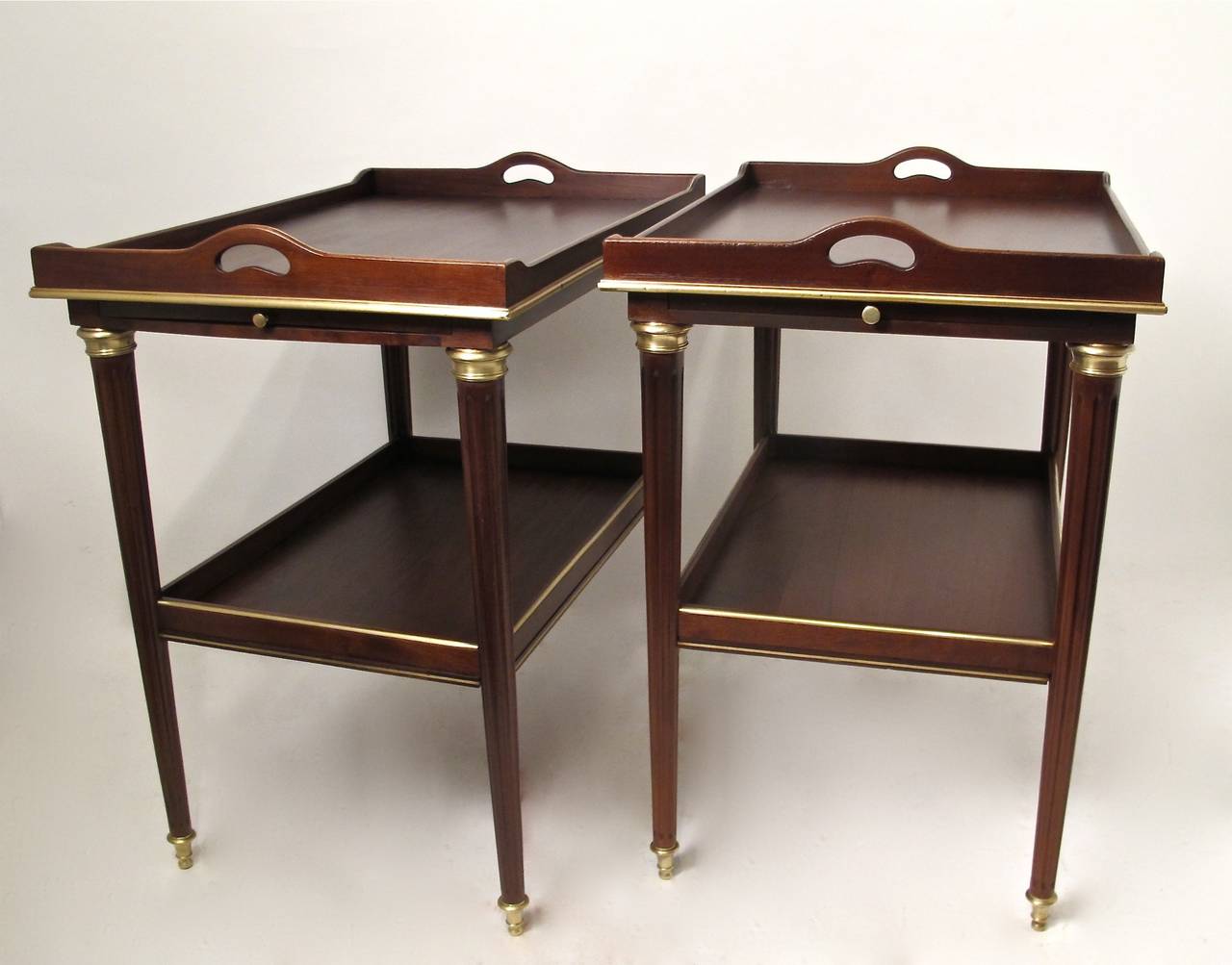 A handsome pair of mahogany tables with glides at either end, a lower shelf, brass inlay and brass mouldings, on tapering legs with brass sabots. Possibly custom French designer made, French, first quarter of the 20th century.