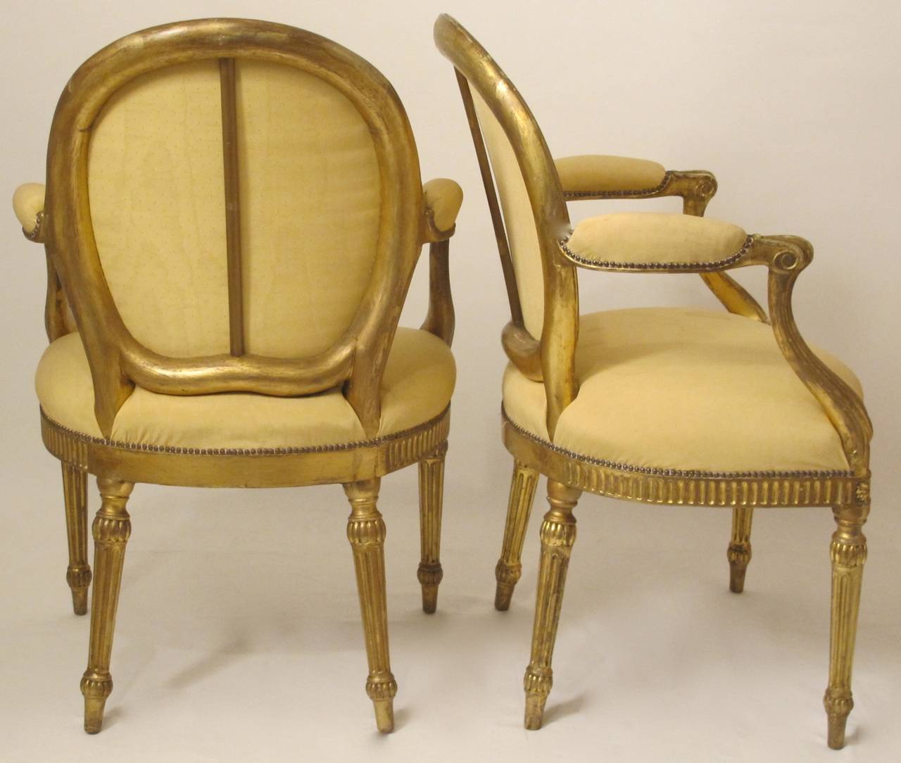 Each with sculpted oval padded back and padded downswept arms and serpentine shaped upholstered seat with fluted rails the center with oval medallion on carved and fluted tapering legs. 
Pegged construction, wear to the original gilding, retouched
