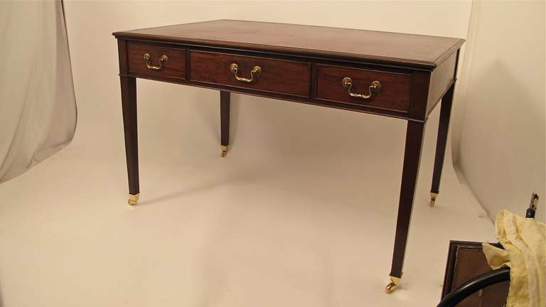 George III style mahogany writing desk with inset burgundy leather with gold tooling surface above two short drawers flanking wider center drawer with brass pulls and castors. False drawers on the front side.  English, Circa 1860.