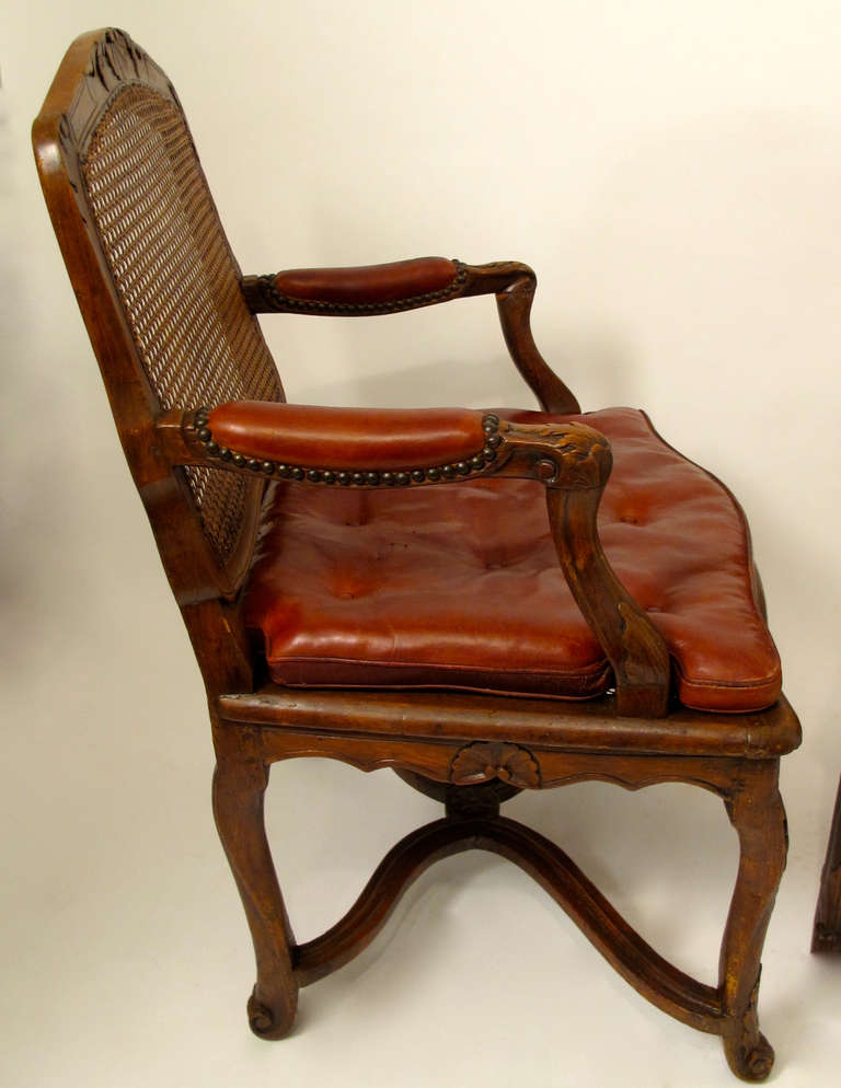 Pair of Regence carved French walnut fauteuils with shell and acanthus carved top rail and leaf carved arms and legs and cane back and seat. The seats upholstered with distressed leather squab button tufted cushions. The hand caning has been
