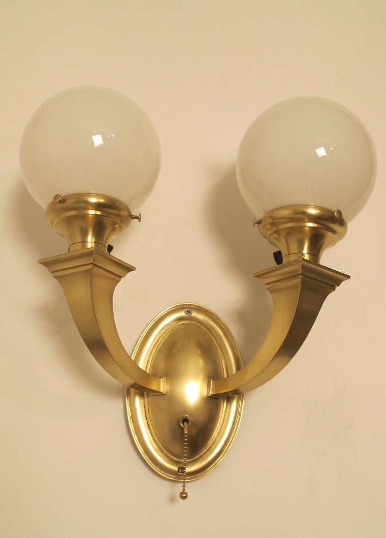 Pair of Brass Two Light Wall Scnces with white globe shades.  Arched arms protruding from an oval brass back plate.  Rewired with there original sockets, each light can be turned on or off by its switched or both can be turned off using the pull