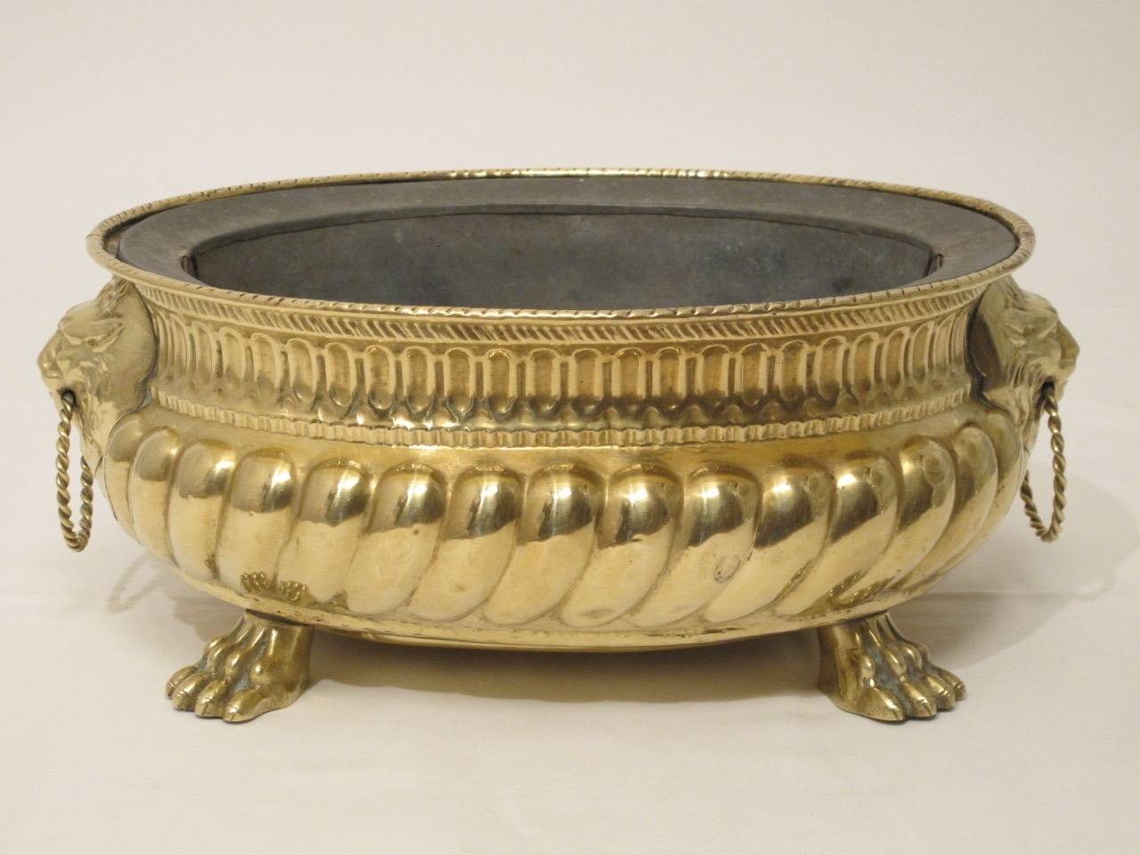 Antique oval brass cache pot with fitted tole liner. Wonderful lion masks holding twisted rope in open mouths. Concave egg and dart design below the broad rim with prominent convex gadrooning supported on lion paw feet.