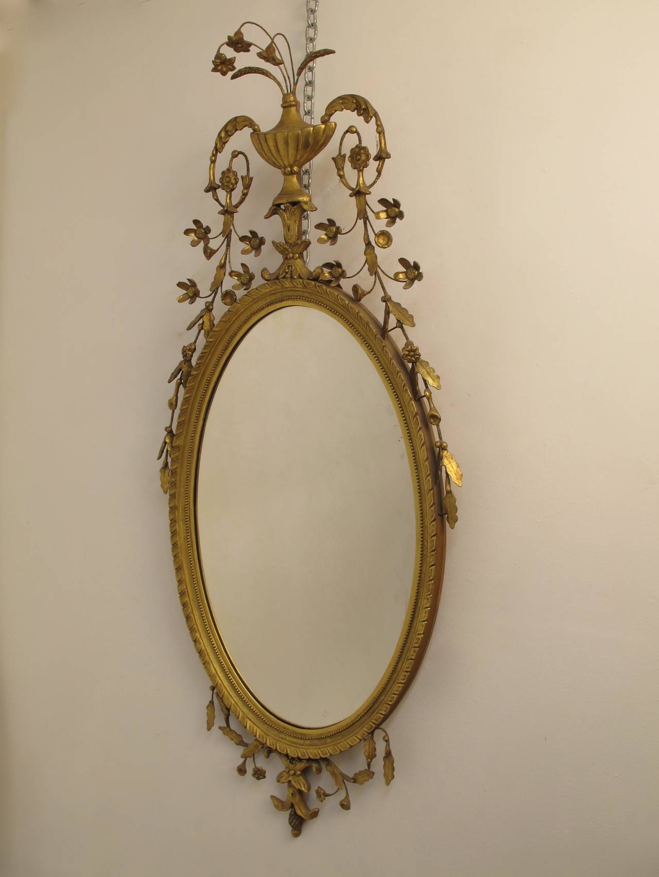 Exubernt Oval Gilt Framed Mirror in the Adams Style. A Central Urn with flowers and hanging swags of leaves from the curled urn handles, leaves and berries trailing partially down the sides.  Made by Kittenger from a Williamsburg 18th. Century