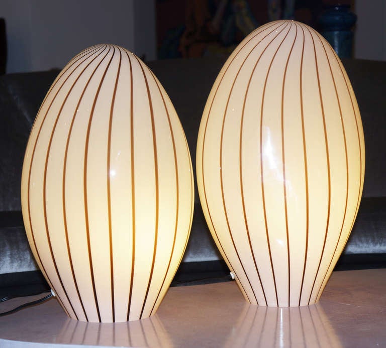Pair of 60's -70's Murano glass oval table lamps made with hand blown glass. Light brown stripes section off the off white surface.
Due to the fact that the lamps are hand blown glass the shapes are somehow different. Each glass contains inside one